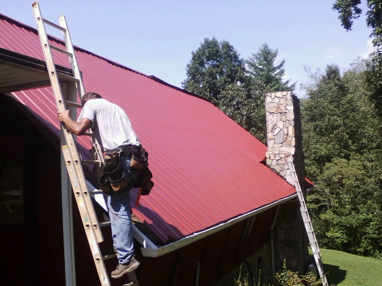 Roofer on a ladder beside a red metal roof and stone chimney — Soffit & Fascia Repair by Roofing Brothers in Illiopolis